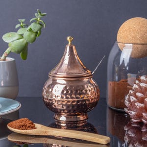 May include: A hammered copper sugar bowl with a lid and a small spoon. The bowl is sitting on a table with a cup and saucer, a plant, and a glass jar with a cork stopper.