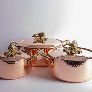 May include: Three copper cooking pots with hammered texture, gold-coloured handles and lid ornaments. The lids are topped with decorative gold-coloured animal figures. The pots are of varying sizes.