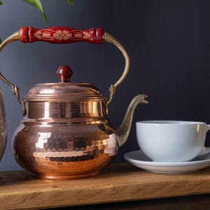 May include: A hammered copper teapot with a red wooden handle and a brass spout. The teapot is sitting on a wooden surface next to a white ceramic teacup and saucer.