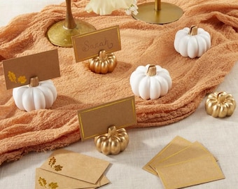 May include: A set of place card holders featuring small white and metallic gold pumpkins. Each pumpkin has a slit at the top to hold rectangular brown kraft paper cards. Some cards are blank, while one features the name Sarah and leaf icons.