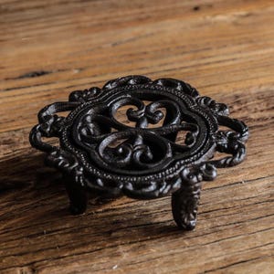 May include: A round, black cast iron trivet featuring an intricate, openwork scroll pattern. The circular piece is supported by three short, sturdy legs and displays a textured, dark finish throughout its detailed metal surface design.