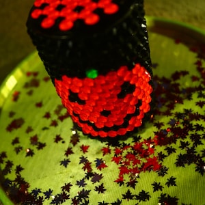 May include: A black and red beaded Halloween-themed candle holder with a smiling jack-o'-lantern face. The holder is sitting on a green tray with red and black confetti.