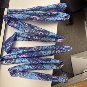May include: Several folded paisley patterned scarves in shades of blue, purple, and white are arranged on a white surface. The scarves have a detailed floral design.