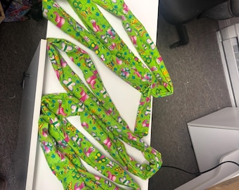 May include: Several green fabric hair wraps with a repeating pattern of pink butterflies and other colorful designs. The wraps are arranged on a white surface. Each wrap has a pink loop at one end.