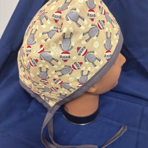 May include: A beige surgical scrub cap featuring a repeating pattern of gray sock monkey illustrations wearing red and white striped hats. The cap has a gray trim and ties.