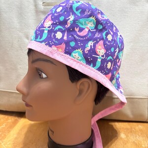 May include: Purple and pink surgical scrub hat with a mermaid and seahorse pattern. The hat has a pink tie and the text "Mermazing" and "Shell Yeah!" is printed on the hat.