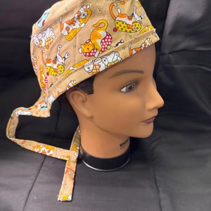 May include: A tan scrub cap featuring a repeating pattern of playful cats in various poses. The cats are in shades of orange, white, and brown, with some wearing colorful accessories. The cap has a tie closure at the back.