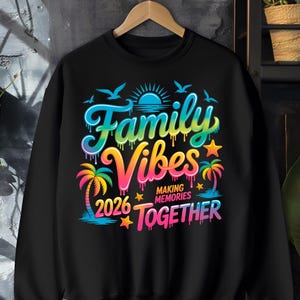May include: Black sweatshirt with a colorful graphic that reads "Family Vibes 2026 Making Memories Together." The design includes palm trees, a sun, birds, and stars, with a rainbow color scheme. The sweatshirt is hanging on a wooden hanger.