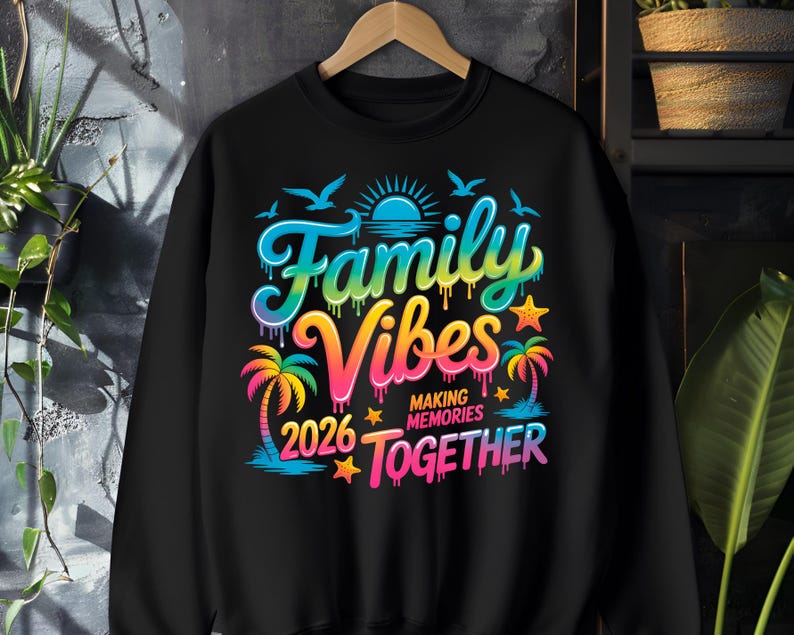 May include: Black sweatshirt with a colorful graphic that reads "Family Vibes 2026 Making Memories Together." The design includes palm trees, a sun, birds, and stars, with a rainbow color scheme. The sweatshirt is hanging on a wooden hanger.