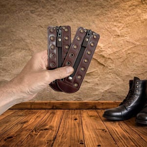 May include: Two dark brown leather boot lace covers featuring a central vertical zipper and two rows of brass-toned metal eyelets along the edges. These rectangular accessories are designed to be attached to the front of footwear.