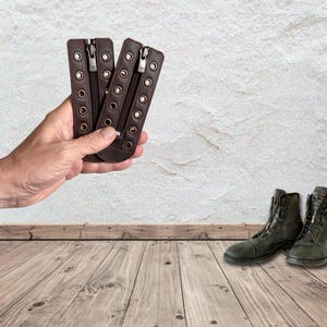 May include: Two dark brown leather boot lace-in zipper inserts featuring a central zipper closure and two vertical rows of metal eyelets along the edges, designed to be attached to the front of lace-up boots for a modified closure system.