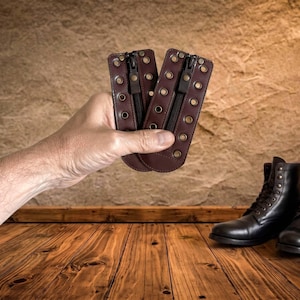 May include: Two dark brown leather boot lace-in zipper inserts featuring a central black zipper, brass-toned metal eyelets along both vertical edges, and a rounded bottom edge, designed to be attached to the front of lace-up boots.