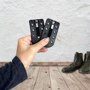 May include: Two black leather boot zipper inserts featuring silver-toned metal eyelets along the edges and a central vertical zipper closure. These rectangular attachments are designed to be laced into footwear to provide a quick-access zipper.