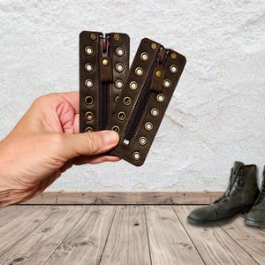 May include: Two rectangular dark brown leather zipper inserts featuring two vertical rows of brass-toned metal eyelets. Each insert has a central zipper closure with a matching leather pull tab and a small brass snap button at the top.