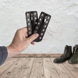 May include: Two black leather boot lace-up inserts featuring a central zipper closure and a series of metallic eyelets along both sides. These rectangular accessories are designed to be attached to footwear to modify the lacing system.