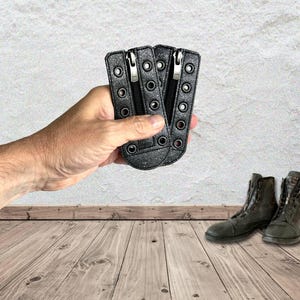 May include: Two black glittery shoe lace zipper inserts featuring a central vertical zipper closure and multiple metal eyelets along the sides for lacing. These rectangular attachments are designed to be placed over the tongue of a boot or shoe.