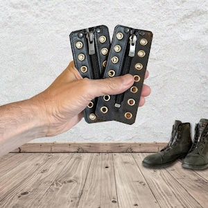 May include: Two black leather zipper pulls with gold eyelets and a silver zipper. The zipper pulls are held in a hand.