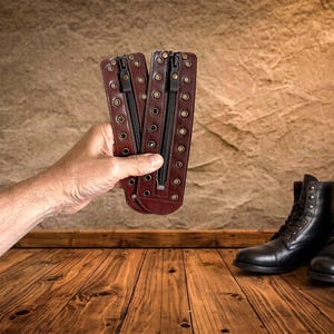 May include: Two dark brown leather boot zipper inserts featuring a central black zipper and two vertical rows of brass-toned metal eyelets along the edges. These rectangular leather panels are designed to be added to lace-up boots for a modified look.