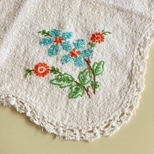 May include: White embroidered cloth with a floral design. The embroidery features blue and orange flowers with green leaves and stems. The edge of the cloth has a decorative, crocheted border.