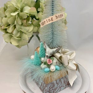 May include: A small, decorative box with a blue and gold floral design. The box is topped with a white ceramic deer figurine, a small blue pine tree, and a white ribbon with the text "spring hope".