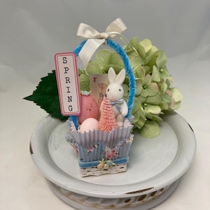 May include: A small blue and white basket with a blue handle, filled with pink Easter eggs and a white bunny figurine. The basket is decorated with flowers and lace. A pink sign with the word "SPRING" is attached to the handle.