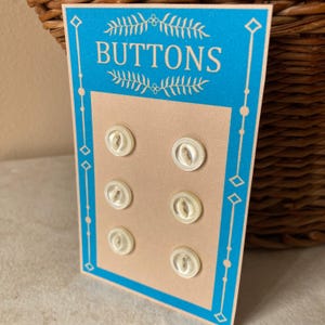 May include: A card of six off-white buttons. The card has a light blue border with the word "BUTTONS" at the top. The buttons are round with a central design and a small hole. The card is tan.