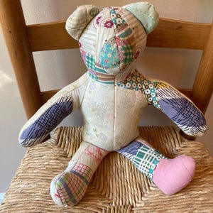 May include: A handmade patchwork teddy bear with a variety of patterned fabrics in pastel colors. The bear has a heart-shaped foot and is sitting on a woven chair. The bear's body is made of floral and striped fabrics.