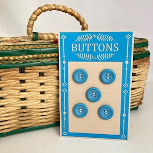 May include: A card of five light blue buttons with two holes each. The card has a blue border with the word "BUTTONS" in white. A woven basket with a handle is in the background.
