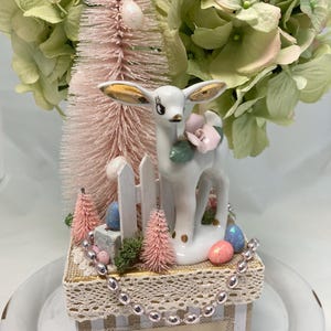 May include: A white porcelain deer figurine with gold accents, holding a pink flower, is surrounded by pink and green miniature trees, pastel Easter eggs, and a white picket fence. The figurine is sitting on a gold and white striped box with a lace trim and the text "easter joy".