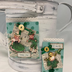 May include: A white distressed metal watering can decorated with St. Patrick's Day themed embellishments. The decorations include images of leprechauns, sheet music, and a bird with a shamrock. The text "happy jig" and "17 March" are visible.
