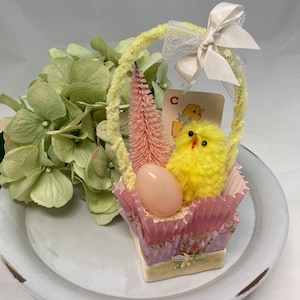May include: A small pink and white Easter basket with a yellow chick, a pink egg, and a pink and white bottle brush tree. The basket is decorated with a white bow and a card that says "C".