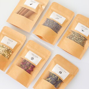May include: Six small brown paper bags with clear plastic windows, each containing different dried herbs and flowers. The bags are labelled with the names of the herbs and flowers: Cinnamon, Lavender, Lotus Leaf, Jasmine, Rose Buds, and Butterfly Pea.