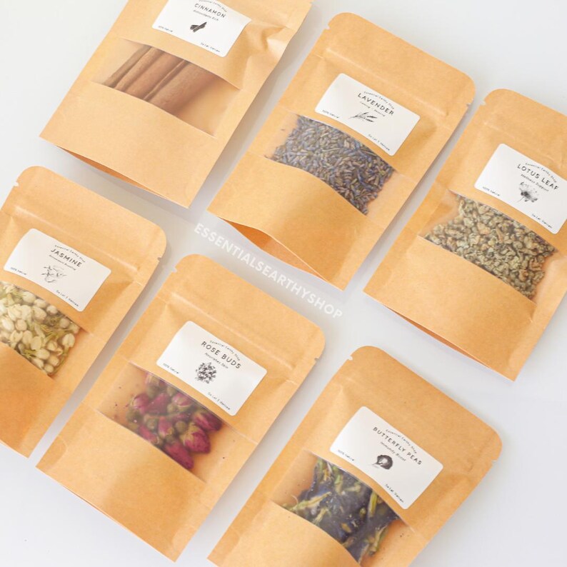 May include: Six small brown paper bags with clear plastic windows, each containing different dried herbs and flowers. The bags are labelled with the names of the herbs and flowers: Cinnamon, Lavender, Lotus Leaf, Jasmine, Rose Buds, and Butterfly Pea.