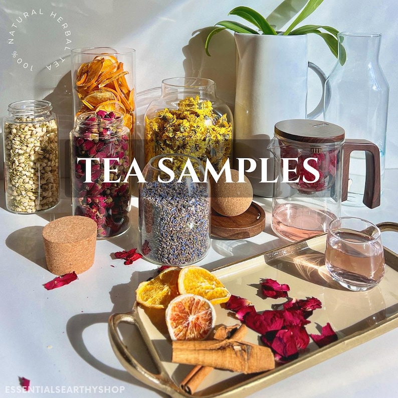 May include: A collection of glass jars filled with various dried herbs and flowers, labelled "TEA SAMPLES". The jars are arranged on a white surface with a wooden tray containing cinnamon sticks, dried orange slices, and red rose petals.