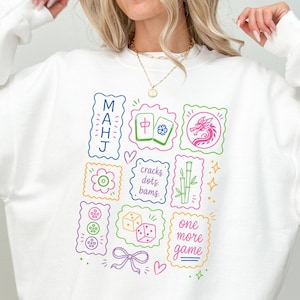 May include: A white long-sleeve sweatshirt featuring a colorful graphic print of Mahjong-themed icons, including tiles, dice, bamboo, and a dragon. The text reads MAHJ, cracks dots bams, and one more game, arranged in a grid of scalloped-edge rectangles.