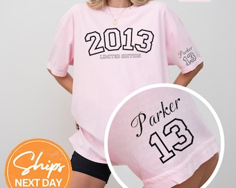 May include: A light pink t-shirt with the year "2013" in black block letters and the words "LIMITED EDITION" below. The sleeve features the name "Parker" and the number "13" in black. The shirt is a casual style.