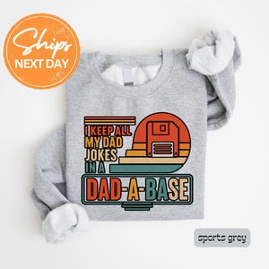 May include: Sports grey sweatshirt with the text "I KEEP ALL MY DAD JOKES IN A DAD-A-BASE" in a retro design. The graphic includes a floppy disk and a sunset color scheme. The sweatshirt is a crew neck.