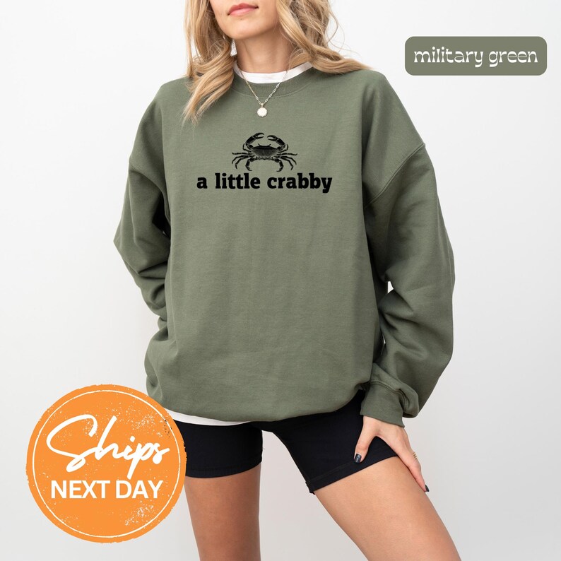 A woman wearing a green sweatshirt with the text "a little crabby" printed on it, standing against a white background.