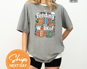 May include: Grey t-shirt with the text "Feeding KIDS is a WORK of HEART" in colorful lettering. The design includes a chef's hat, hearts, and cooking utensils. The shirt is a relaxed fit.
