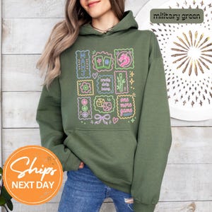 May include: A military green hooded sweatshirt featuring a graphic print of neon-style Mahjong tiles, a dragon, bamboo, dice, and the text "MAHJ", "cracks dots bams", and "one more game" in various colors including pink, blue, and yellow.