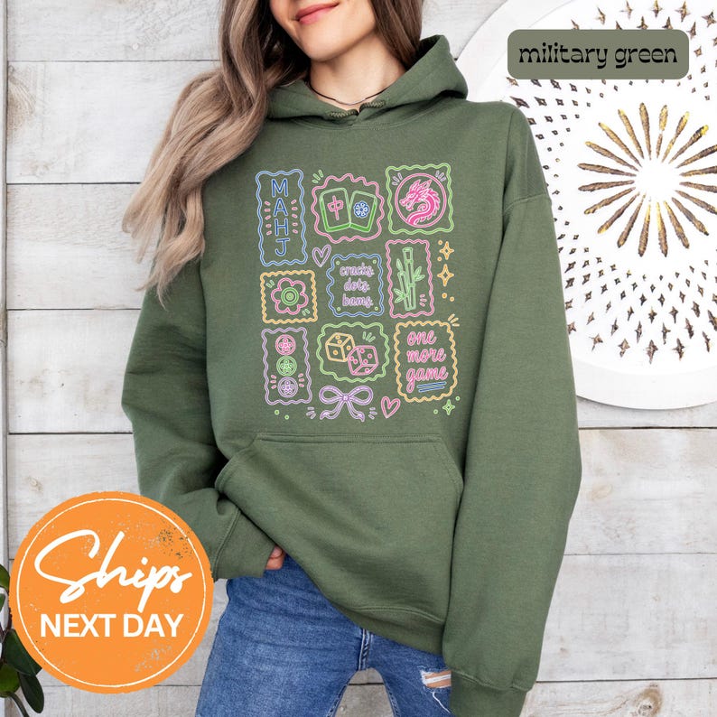 May include: A military green hooded sweatshirt featuring a graphic print of neon-style Mahjong tiles, a dragon, bamboo, dice, and the text "MAHJ", "cracks dots bams", and "one more game" in various colors including pink, blue, and yellow.