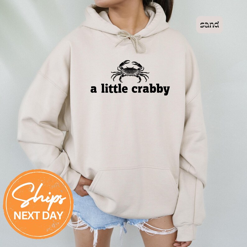 A person wearing a beige hoodie with a black crab design and the text "a little crabby" printed on it.