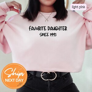May include: Light pink sweatshirt with the text "FAVORITE DAUGHTER SINCE 1993" in black. The sweatshirt has long sleeves and a crew neck. The image also includes a black belt with a silver buckle.