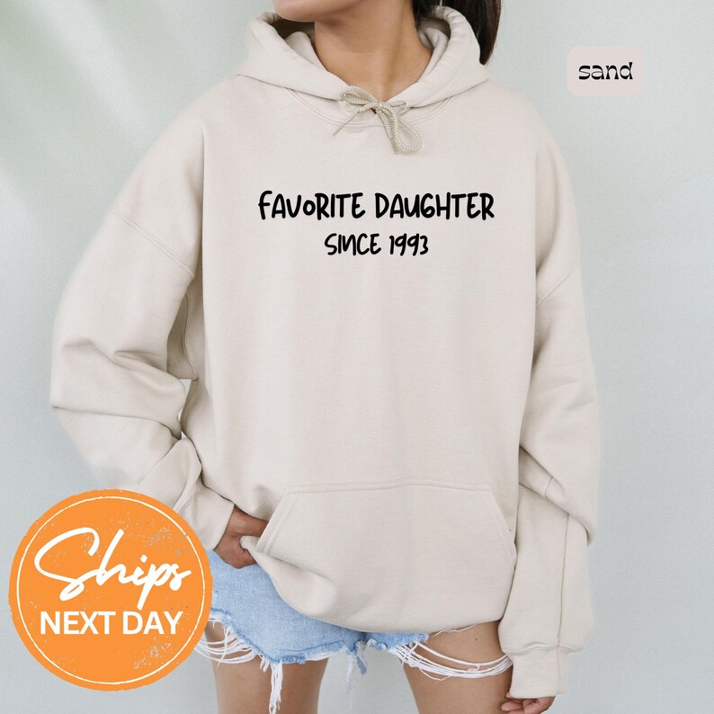 May include: Sand-colored hoodie with the text "FAVORITE DAUGHTER SINCE 1993" in black. The hoodie has a drawstring hood and a front pocket. The image also includes a "Ships Next Day" graphic.