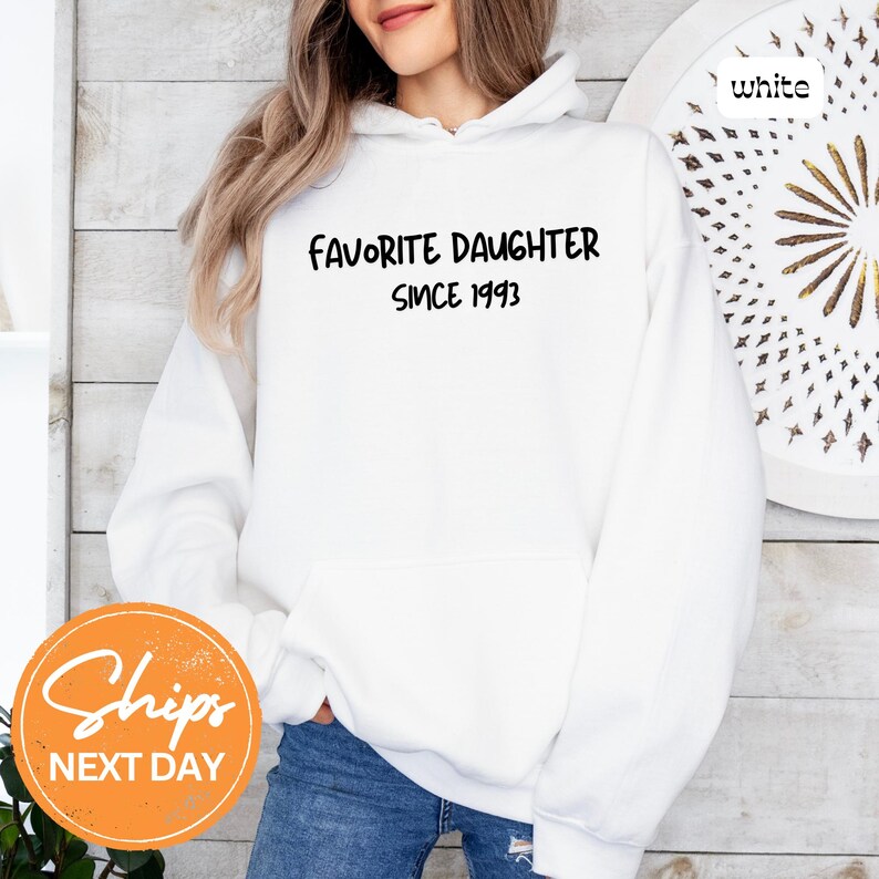 May include: White hoodie with the text "FAVORITE DAUGHTER SINCE 1993" in black. The hoodie has a front pocket and a hood. The image also includes a round orange sticker with the text "Ships NEXT DAY".