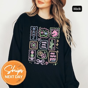 May include: A black long-sleeve sweatshirt featuring a neon-style graphic print with Mahjong-themed illustrations, including tiles, a dragon, bamboo, dice, and text that reads "MAHJ", "cracks dots bams", and "one more game" in vibrant colors.