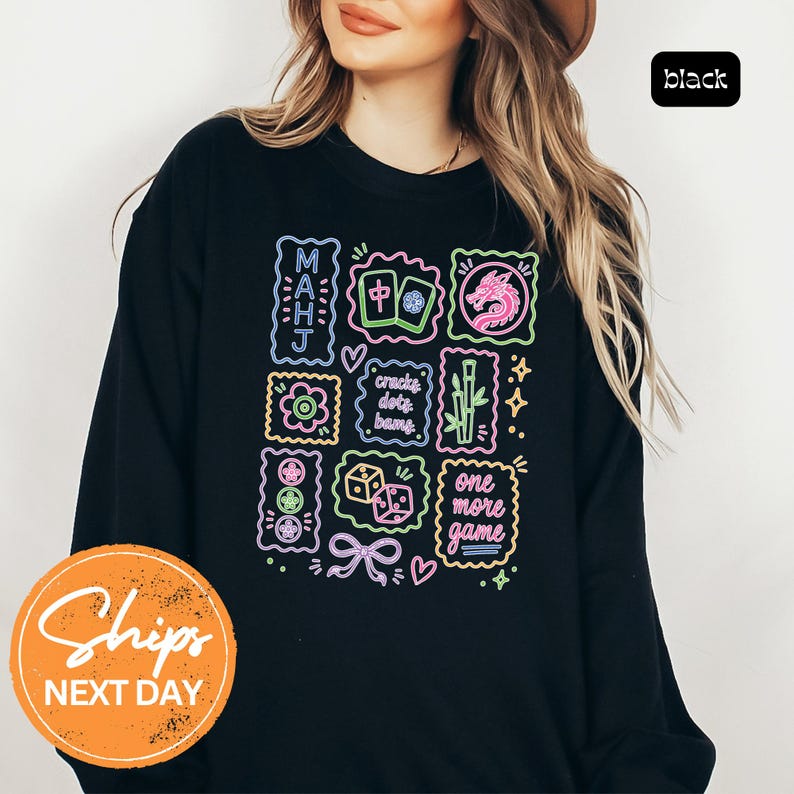 May include: A black long-sleeve sweatshirt featuring a neon-style graphic print with Mahjong-themed illustrations, including tiles, a dragon, bamboo, dice, and text that reads "MAHJ", "cracks dots bams", and "one more game" in vibrant colors.