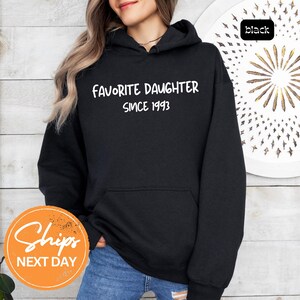 May include: Black hooded sweatshirt with the text "FAVORITE DAUGHTER SINCE 1993" in white. The sweatshirt has a front pocket and a drawstring hood. The word "black" is in a small black box in the upper right corner.