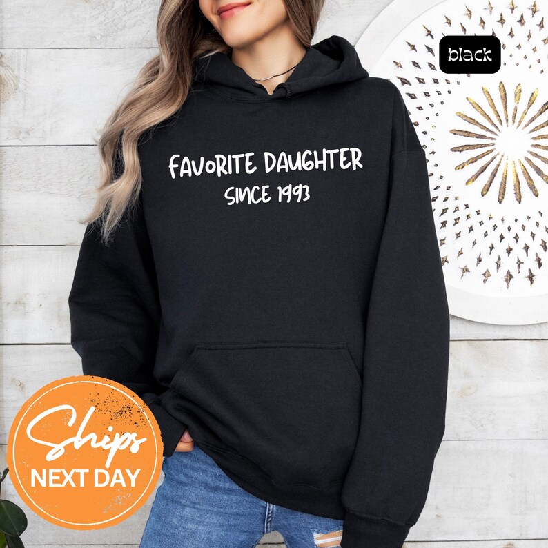 May include: Black hooded sweatshirt with the text "FAVORITE DAUGHTER SINCE 1993" in white. The sweatshirt has a front pocket and a drawstring hood. The word "black" is in a small black box in the upper right corner.