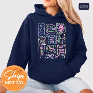 May include: A navy blue hooded sweatshirt featuring a colorful neon-style graphic print on the front. The design includes Mahjong tiles, a dragon, bamboo, dice, and text that reads MAHJ, cracks dots bams, and one more game in various colors.